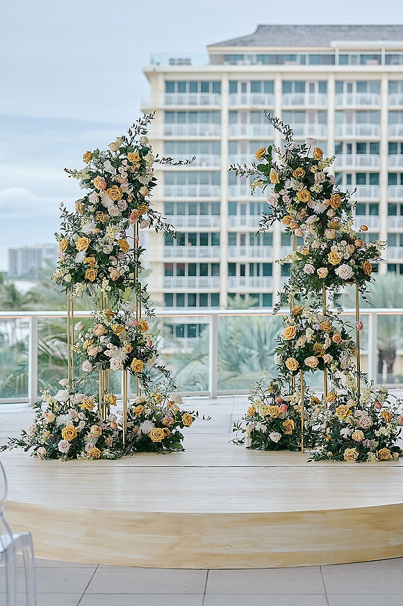 Wedding ceremony backdrop with floral ceremony backdrop pillars of roses and greenery on gold stands atop a terrace platform with ocean view