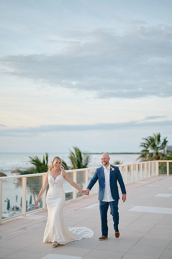 Couple portrait of bride and groom holding hands, walking on a rooftop terrace with ocean view, palm trees, and her dress train flowing