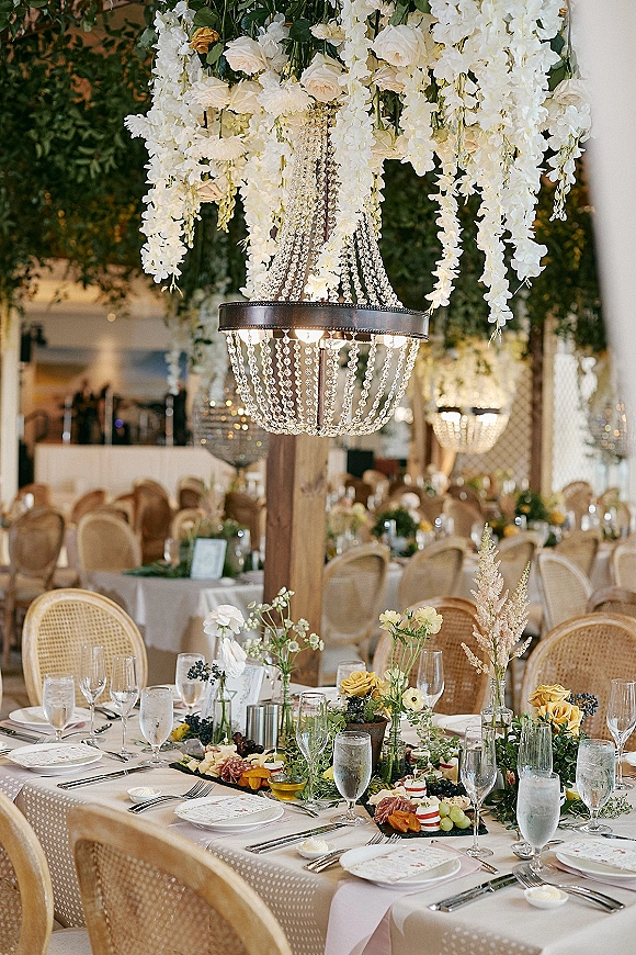 Reception tablescape with crystal chandelier centerpiece, hanging white florals, taper candles, and rattan chairs in a banquet hall