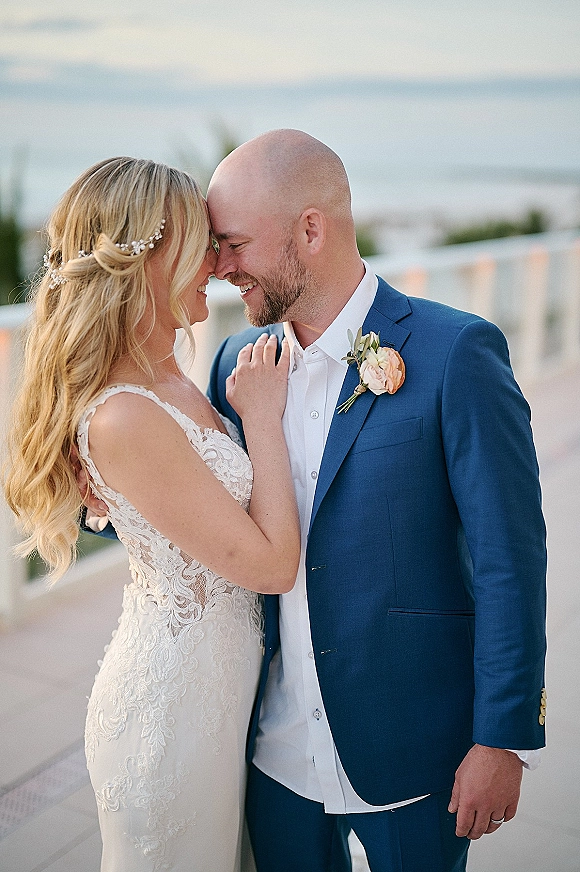 Couple portrait on a beach wedding couple touching foreheads, bride in lace dress with hair vine and groom in blue suit by the ocean