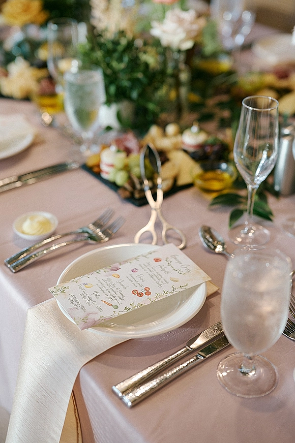 Reception tablescape with wedding place setting featuring a menu card on white plates, silver flatware, glassware, and greenery centerpiece on pink linen