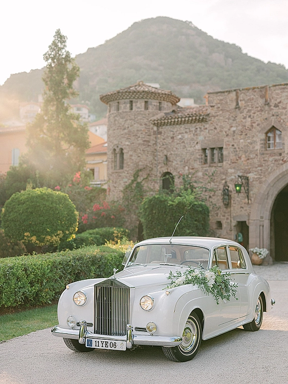Wedding getaway car, a vintage white car with white flowers and greenery garland, parked by a stone archway with mountains beyond