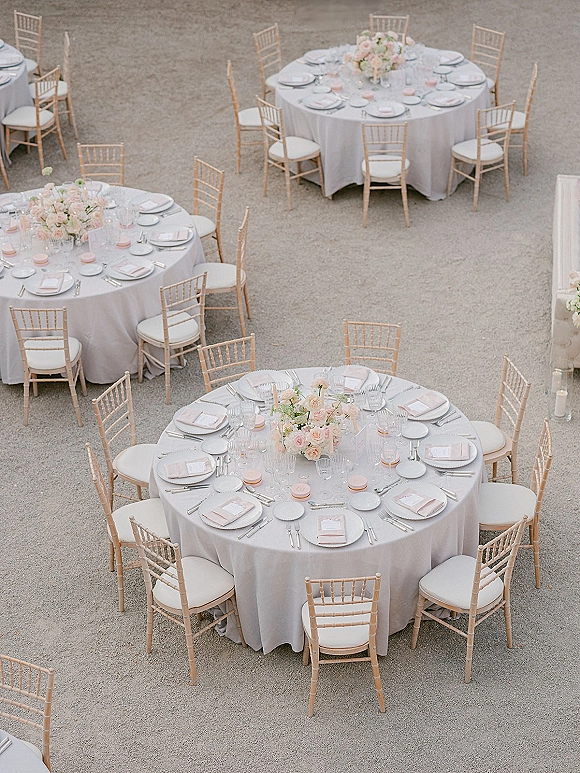 Wedding reception tablescape with round wedding reception tables dressed in white linens, blush floral centerpieces, and votive candles in a gravel courtyard