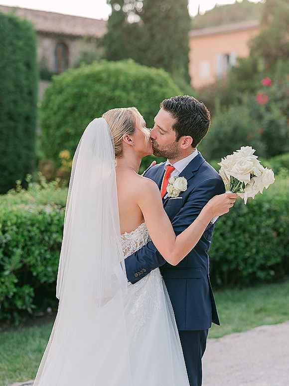 Wedding kiss portrait of bride and groom kissing, her long veil and lace dress flowing as she holds a white rose bouquet in a villa garden