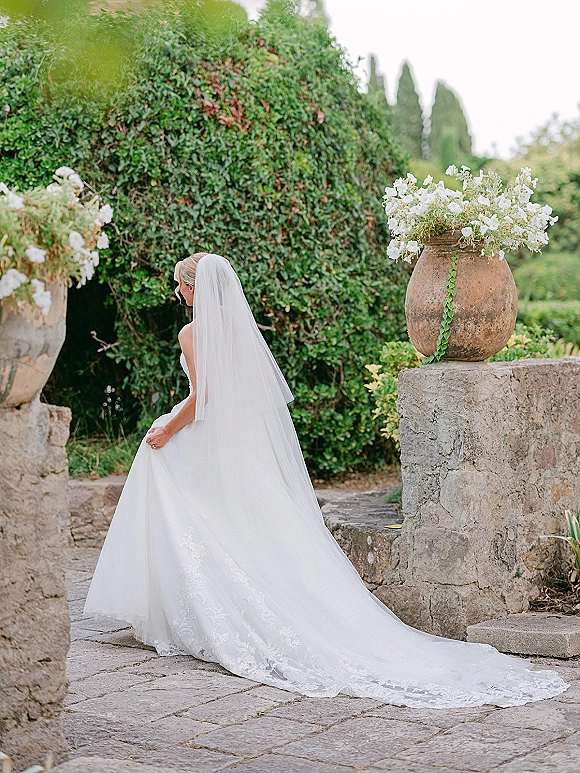 Bridal portrait of bride back view in a strapless gown with cathedral veil and lace train, walking up stone courtyard steps past floral urns