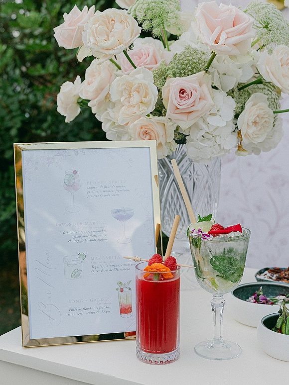 Wedding cocktail bar with signature drink menu in a gold frame, pastel florals, and garnished cocktails on a white table in greenery