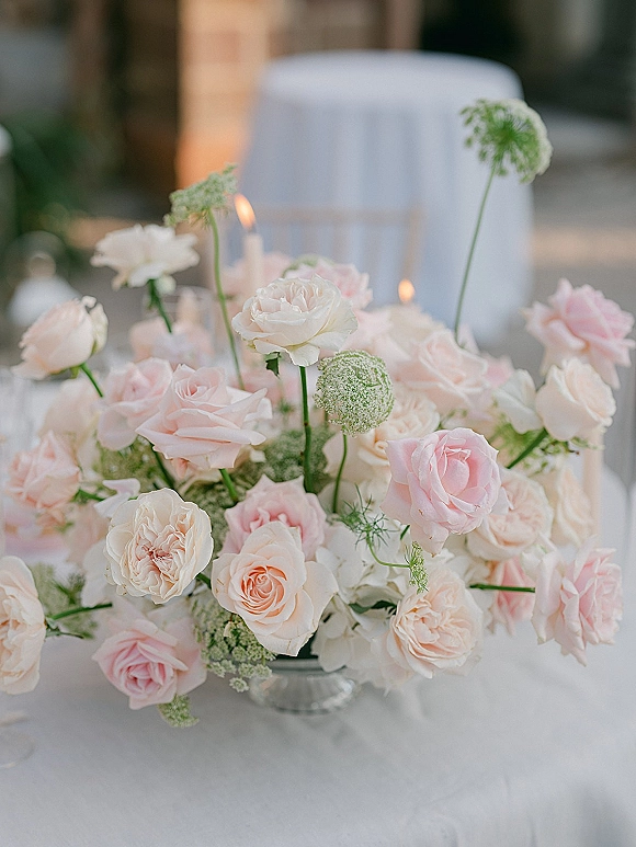Wedding centerpiece with blush rose centerpiece of ivory and soft pink roses, greenery in a glass compote, taper candles on a white tablecloth