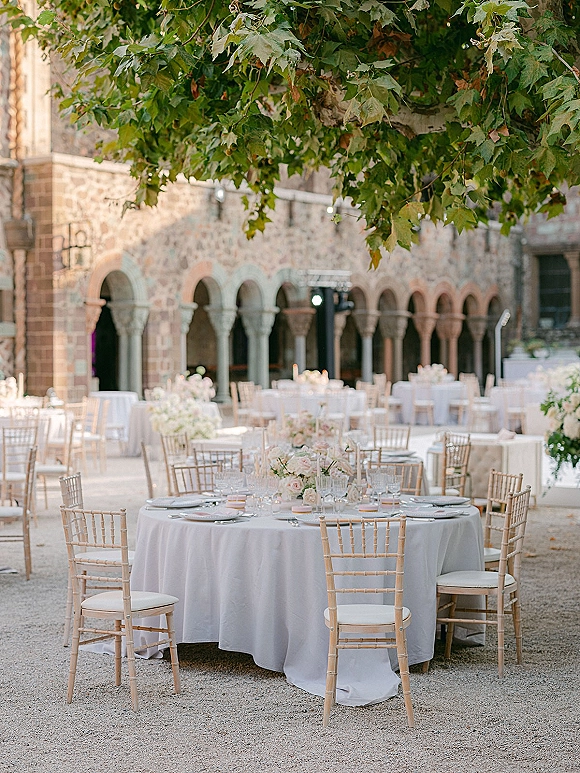 Outdoor reception setup with round tables, white linens, floral centerpieces, and gold chiavari chairs under string lights in a stone courtyard