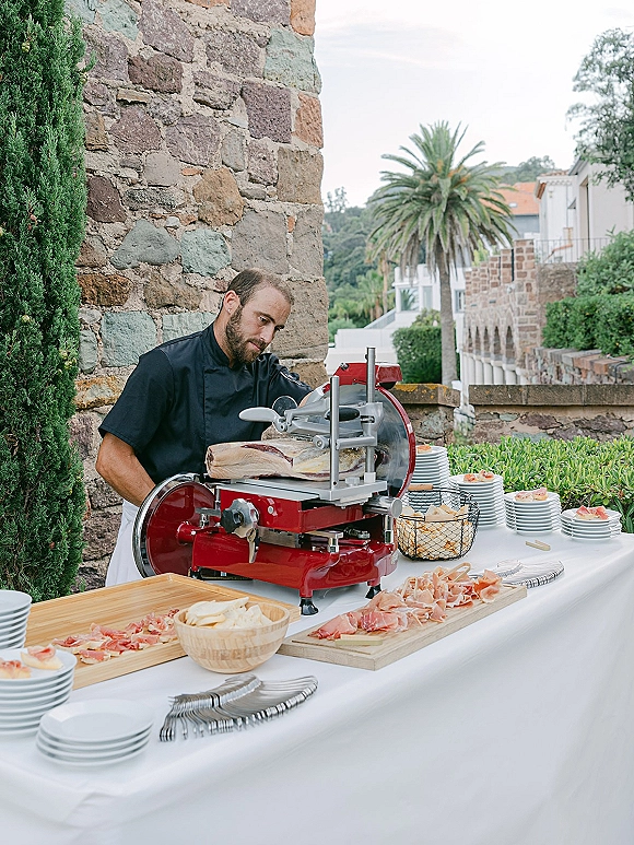 Wedding cocktail hour buffet with cocktail hour charcuterie, chef slicing cured ham beside cheeses and breadsticks on an outdoor terrace stone wall backdrop