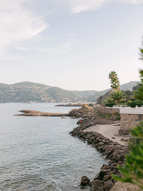 Coastal landscape with rocky shoreline view along a seaside stone wall accent, with palm trees, boats in the bay, and hills beyond