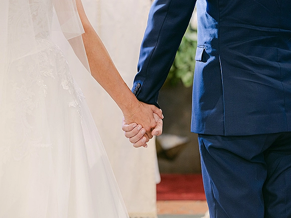Couple holding hands, wedding handholding photo from behind with bridal veil, tulle dress, navy suit, and bracelet on a green walkway