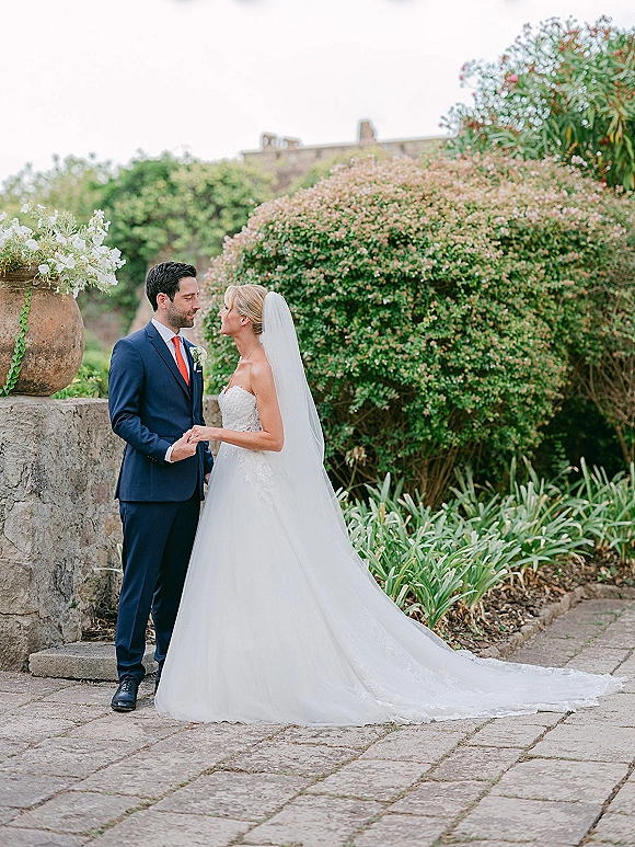 Couple portrait of bride and groom holding hands, face to face in a stone courtyard with greenery, her cathedral veil flowing behind