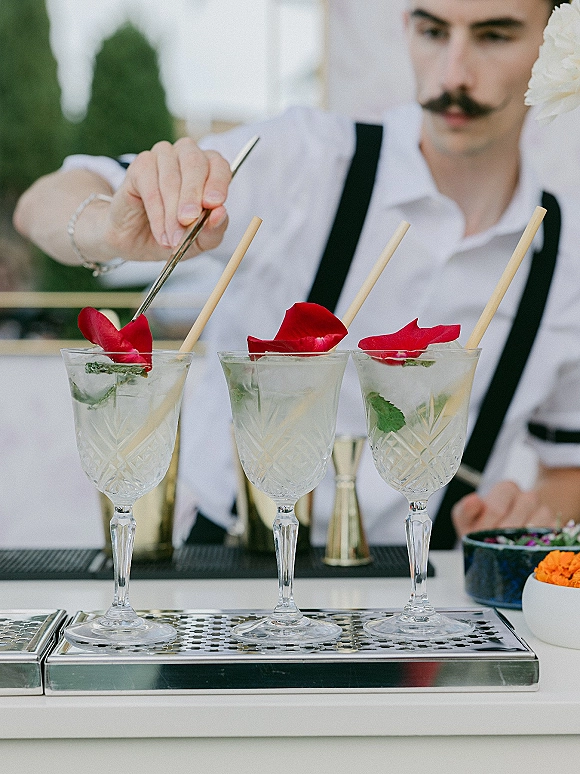 Signature wedding cocktails in coupe glasses with mint and red rose petal garnishes as a bartender preps them at an outdoor bar counter.