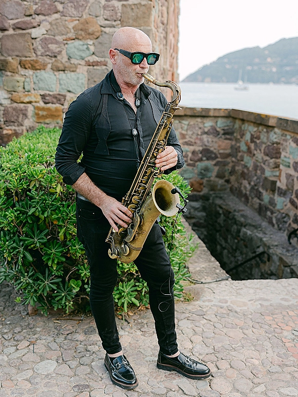 Wedding saxophonist playing live saxophone wedding music in sunglasses and black outfit on a stone terrace by the water with sailboats
