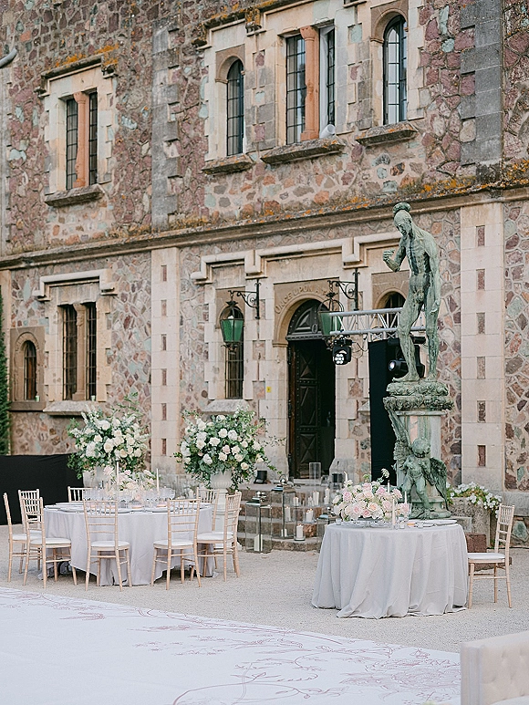 Reception tablescape with round tables in white linens, lush floral centerpieces and taper candles in a stone manor courtyard with arched doorway