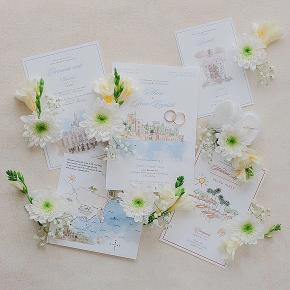 Wedding invitation suite with illustrated wedding invitations, RSVP and map cards, wedding rings, and white flowers on a beige fabric backdrop