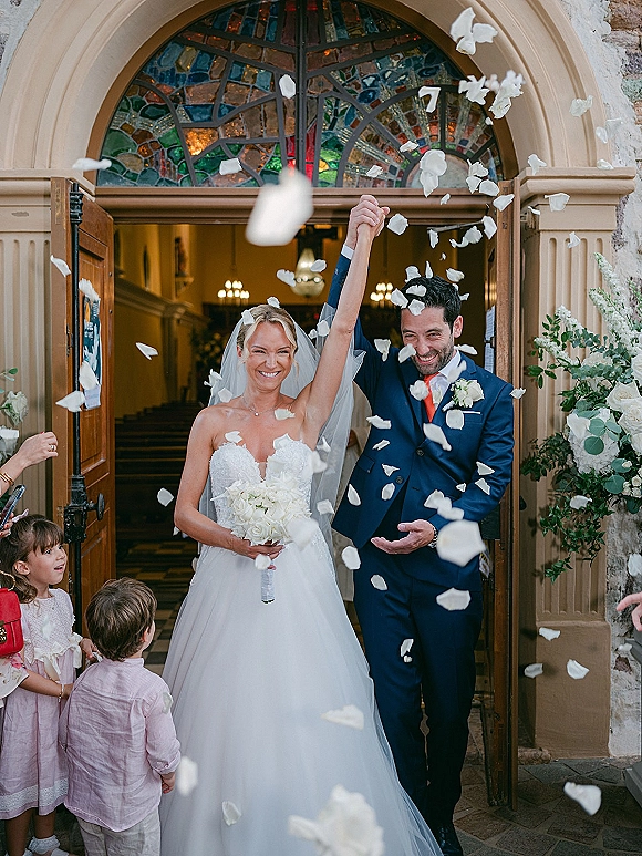 Wedding recessional as bride and groom walk out a church doorway under a rose petal toss, holding hands high, bouquet and veil flowing