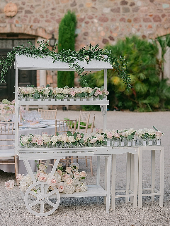 Wedding flower cart with blush and white roses, eucalyptus garland, and glass vases beside a round table in a stone-walled courtyard patio