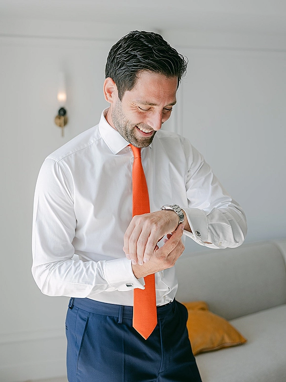 Groom getting ready, adjusting watch in a white dress shirt and orange tie, with cufflinks, navy pants, and sofa behind him