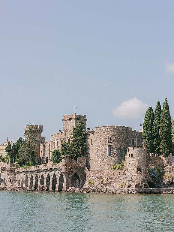 Castle wedding venue on the waterfront, a historic stone castle with turrets and arched seawall beneath blue sky and trees