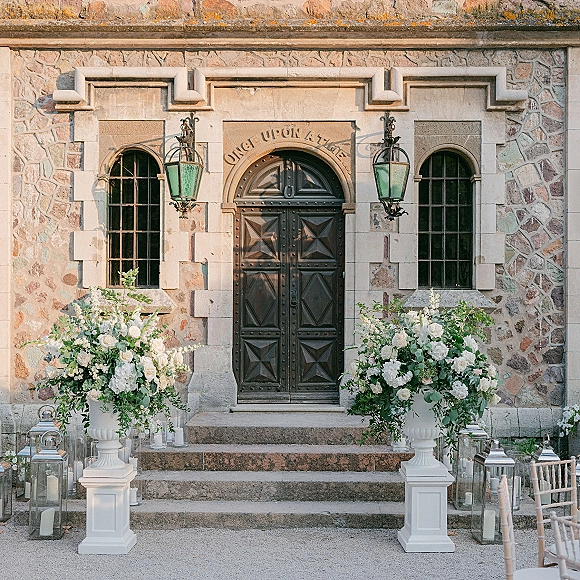 Ceremony entrance decor with wedding doorway florals in white and greenery on urn pedestals, lanterns, and candles by stone steps and doors