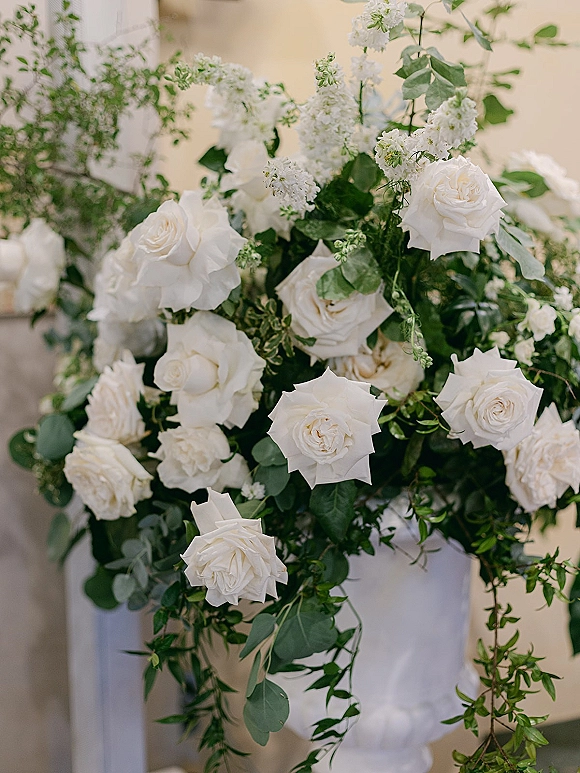 Wedding floral arrangement of white roses and lilac with eucalyptus accents, cascading greenery vines against a neutral wall in soft indoor light