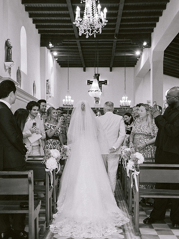 Wedding processional with bride walking down aisle in a long veil and gown train, guests filming beside pews under church chandelier and cross