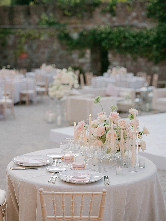 Reception tablescape with blush roses and white florals, tapered candles, crystal glassware, and pink napkins on an outdoor patio by a stone wall