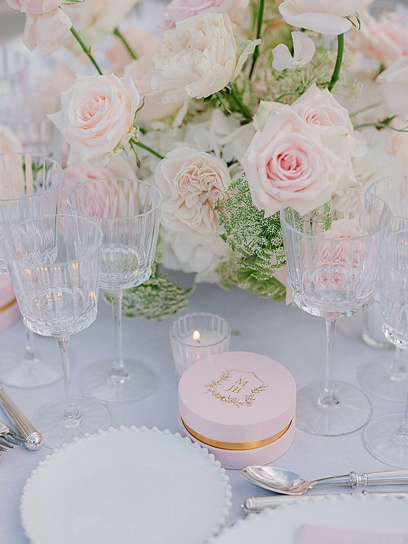 Reception tablescape with a wedding table centerpiece of blush roses and white florals, crystal wine glasses, votive candle, and monogram favor box