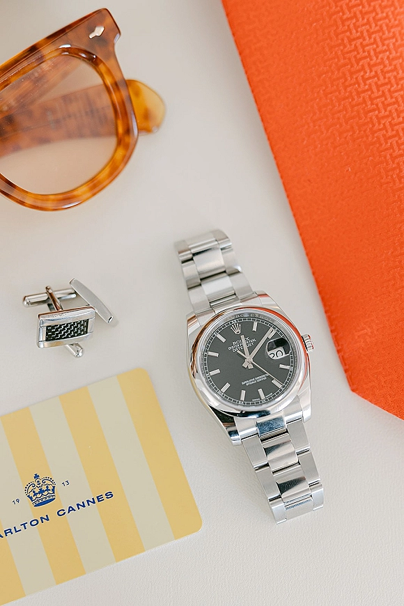 Groom accessories flatlay featuring wedding cufflinks, wristwatch, sunglasses, hotel key card, and an orange pocket square on white tabletop