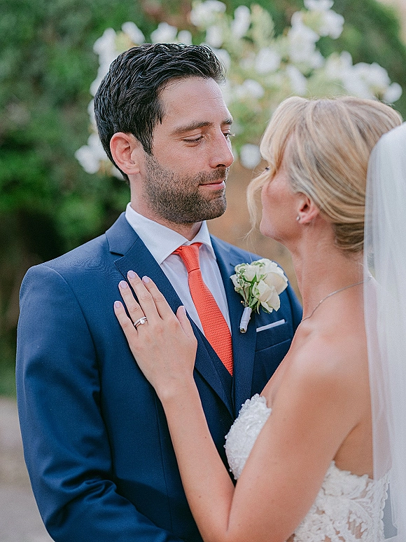 Couple portrait of bride and groom close up, holding each other as she touches his lapel, veil and ring visible amid greenery and white flowers