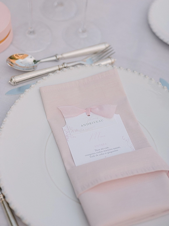 Wedding place setting with blush napkin, menu card tied with a ribbon bow, silver flatware and wine glass on a white tablecloth