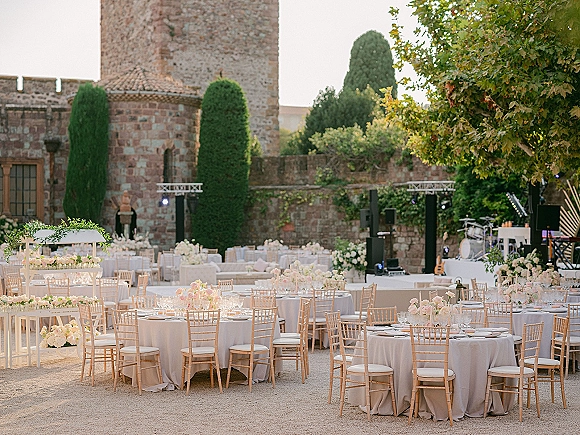Reception tablescape at an outdoor wedding reception with round white-linen tables, floral centerpieces, candles, and a band stage in a castle courtyard