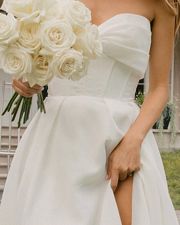 Bridal bouquet of white roses held against a strapless wedding gown, engagement ring visible, with outdoor greenery and steps behind