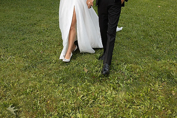 Couple walking hand in hand, bride’s thigh-high slit wedding dress and heels beside groom in a tuxedo on a grass lawn