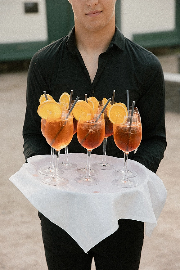 Wedding cocktail service with a waiter in a black button-up carrying stemmed spritz glasses with orange garnish on an outdoor walkway