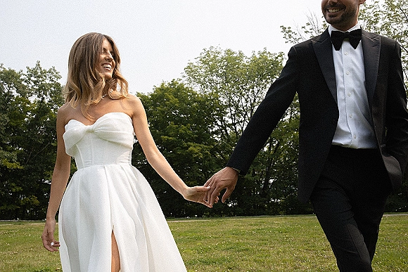 Couple portrait of bride and groom holding hands, laughing in a grassy field under open sky, her strapless slit gown and his black tuxedo bow tie