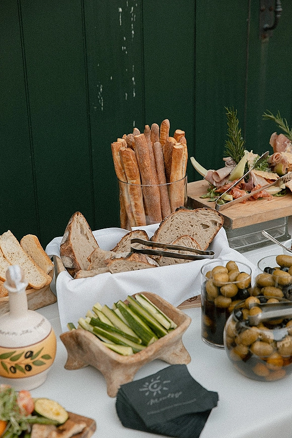 Wedding appetizer table with cocktail hour food table spread of baguettes, sliced bread, olive jars, and charcuterie on wooden boards by a dark paneled wall
