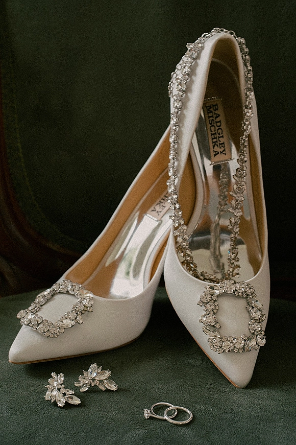 Bridal shoes in a wedding shoes flatlay, white satin heels with crystal buckle, rhinestone straps, rings and earrings on green velvet chair