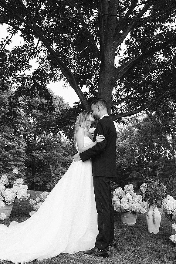 Wedding couple portrait in black and white, bride and groom hugging with foreheads touching under a large tree beside potted hydrangeas