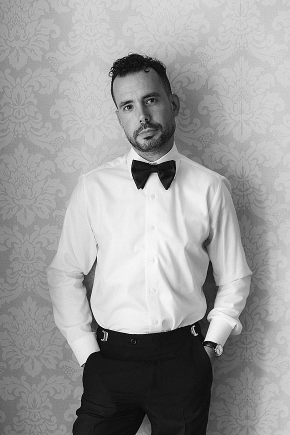 Groom portrait in a groom bow tie, white dress shirt and black pants, wearing a wristwatch against patterned wallpaper backdrop