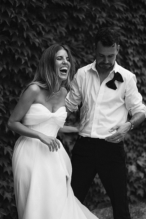 Couple portrait in a black and white wedding photo, bride laughing while holding groom’s arm beside an ivy wall greenery backdrop