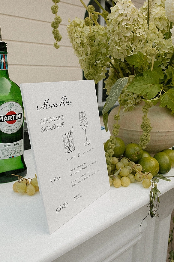 Bar menu sign with wedding bar menu calligraphy, cocktail illustrations, vermouth bottle, and hydrangea vase on a bar counter by a white paneled wall