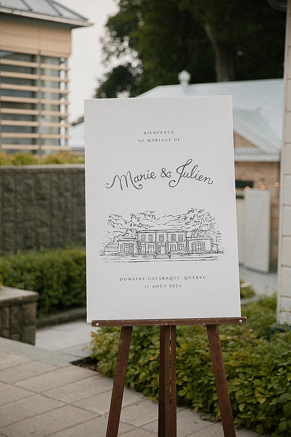 Wedding welcome sign with calligraphy lettering on an easel, featuring a venue illustration along an outdoor walkway with hedges and stone wall