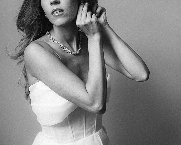Bridal portrait in black and white of a bride in a strapless wedding dress adjusting her stud earring, crystal necklace on a plain studio backdrop