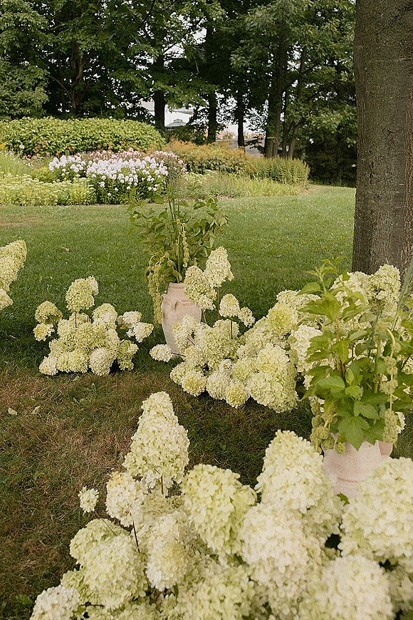 Ceremony aisle florals with hydrangea aisle flowers arranged in ceramic vases, lining a grassy garden path with trees and shrubs beyond