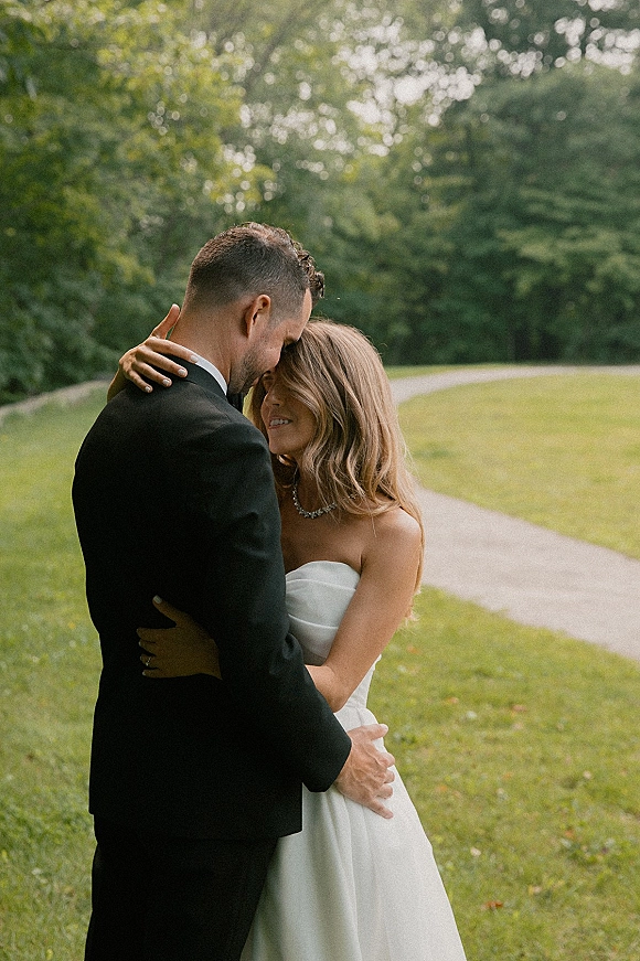 Couple portrait of bride and groom embrace, touching foreheads on a park path with green lawn and trees, bride in strapless gown