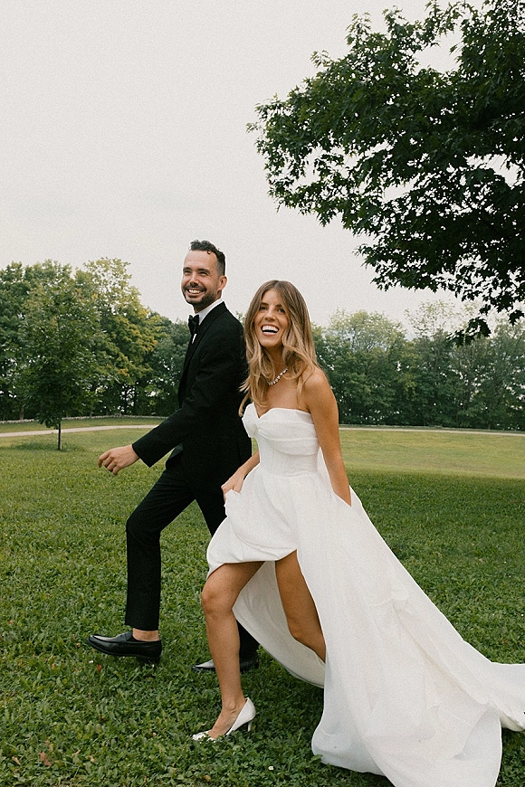 Couple portrait of bride and groom walking, laughing on a grassy park lawn under overcast sky, she wears a strapless high-slit gown and heels