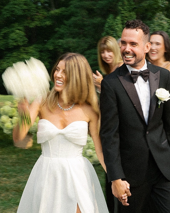 Recessional moment as bride and groom recessional holding hands, bride waving bouquet beside guests amid lush garden greenery outdoors