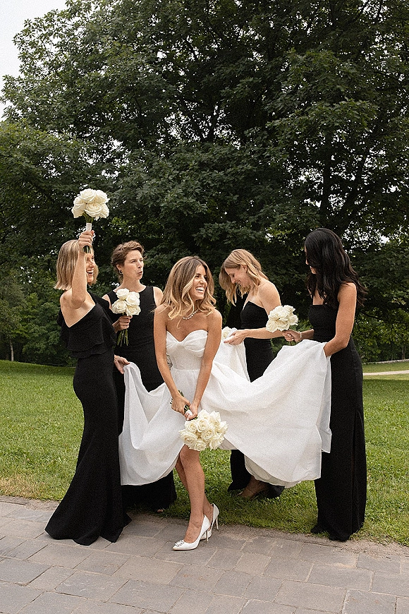 Bride with bridesmaids laughing as bridesmaids holding dress train, all holding white rose bouquets on a lawn by trees and stone walkway